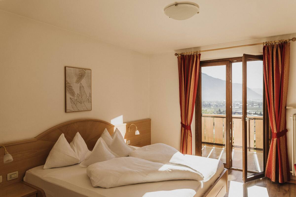 A hotel room with a bed, a nightstand, a lamp, a painting, and a balcony with curtains.
