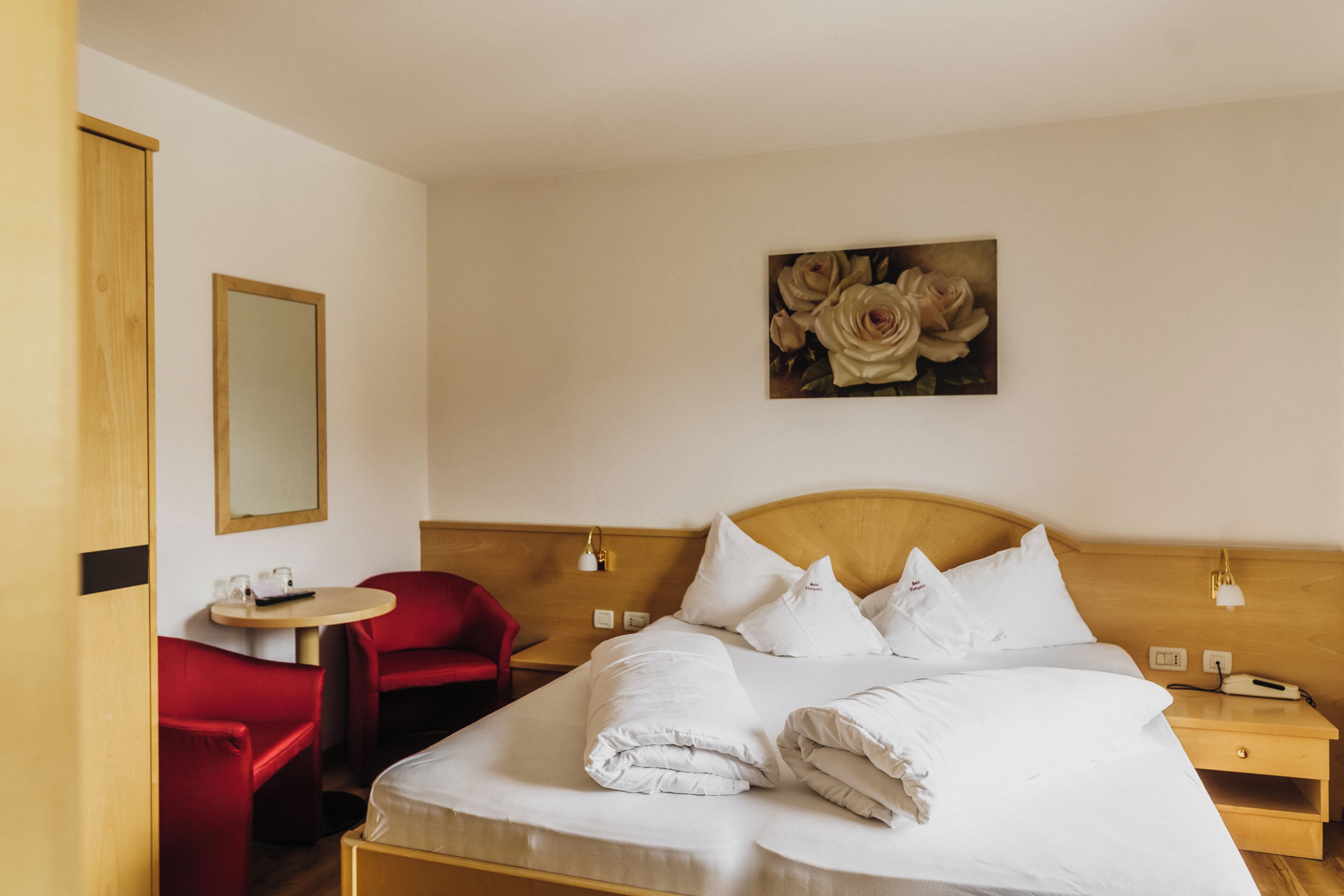 A hotel room with a bed, a red chair, a small table, and a painting on the wall.