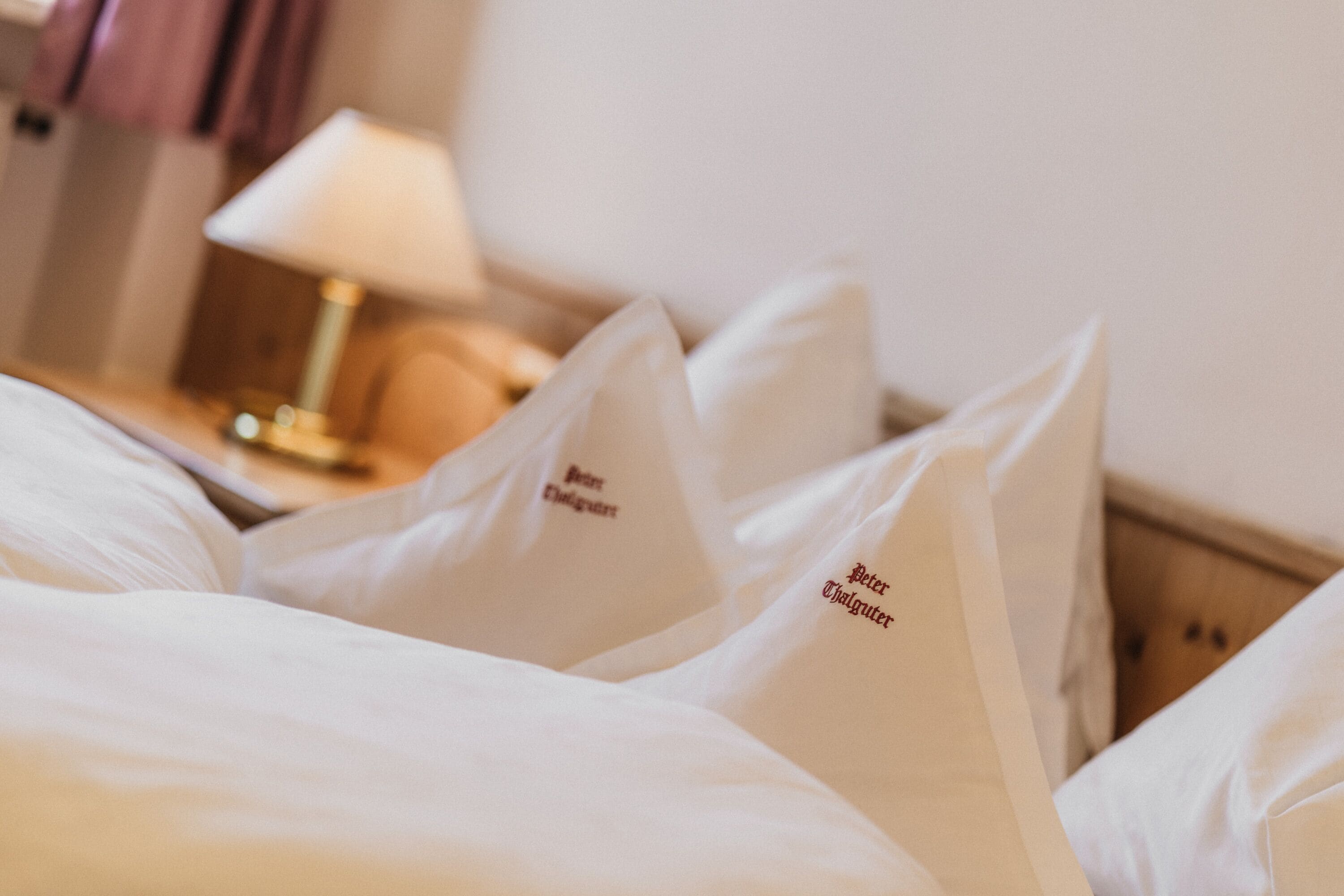 A hotel room with neatly made beds, pillows with embroidered text, a bedside lamp, and a wooden headboard.
