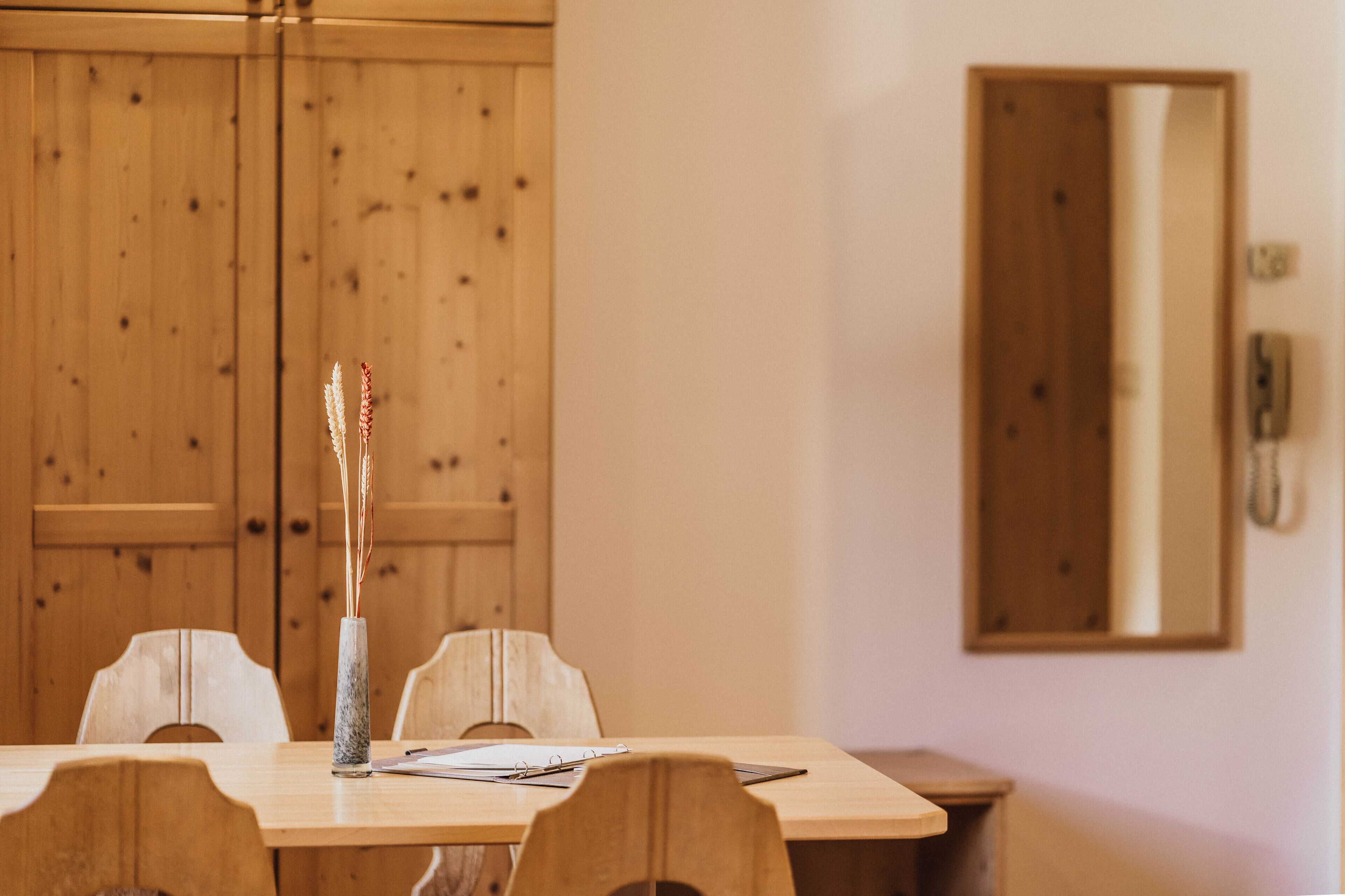 A wooden dining table with chairs, a vase with dried plants, and a mirror on the wall.