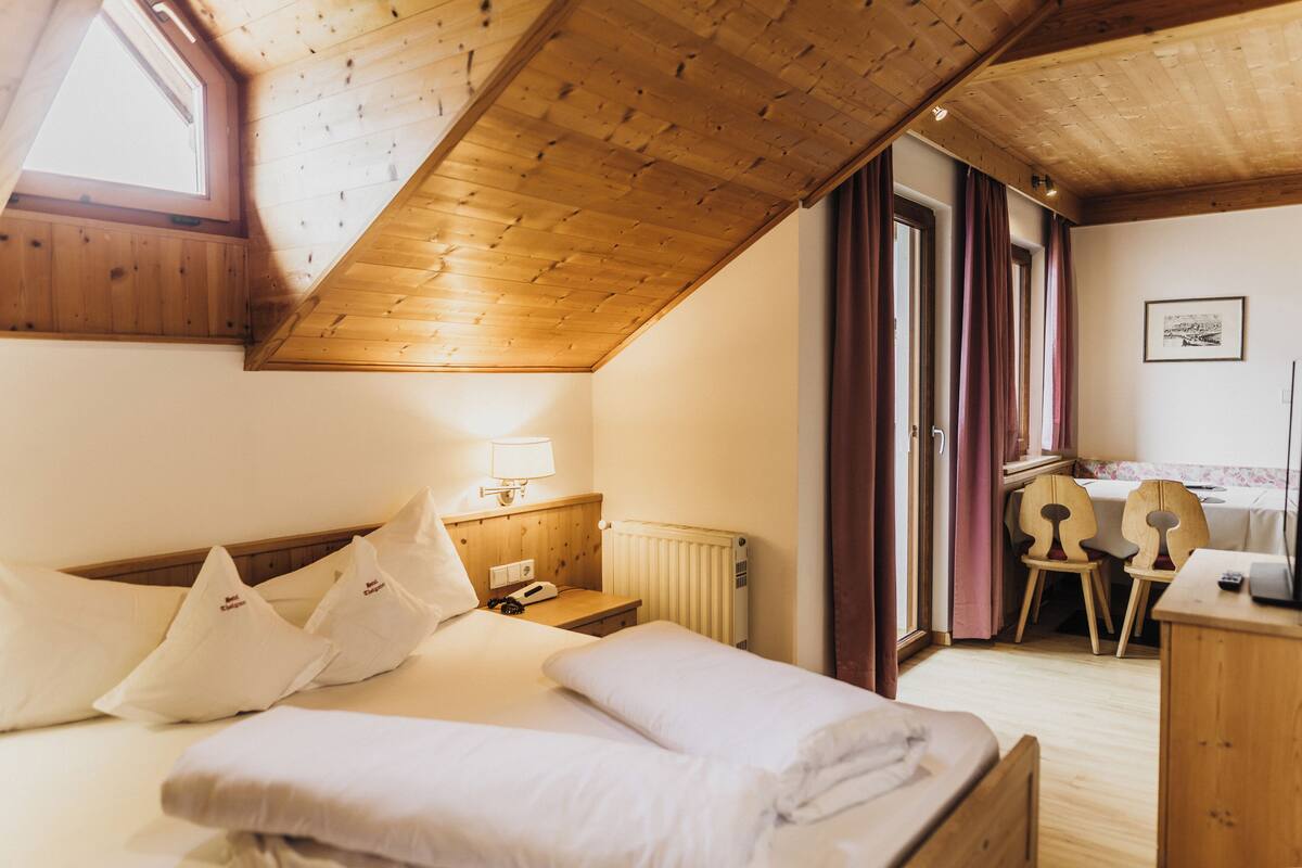 A cozy bedroom with a sloped ceiling, a bed with white linens, a bedside table with a lamp, and a view of a dining area through an open door.