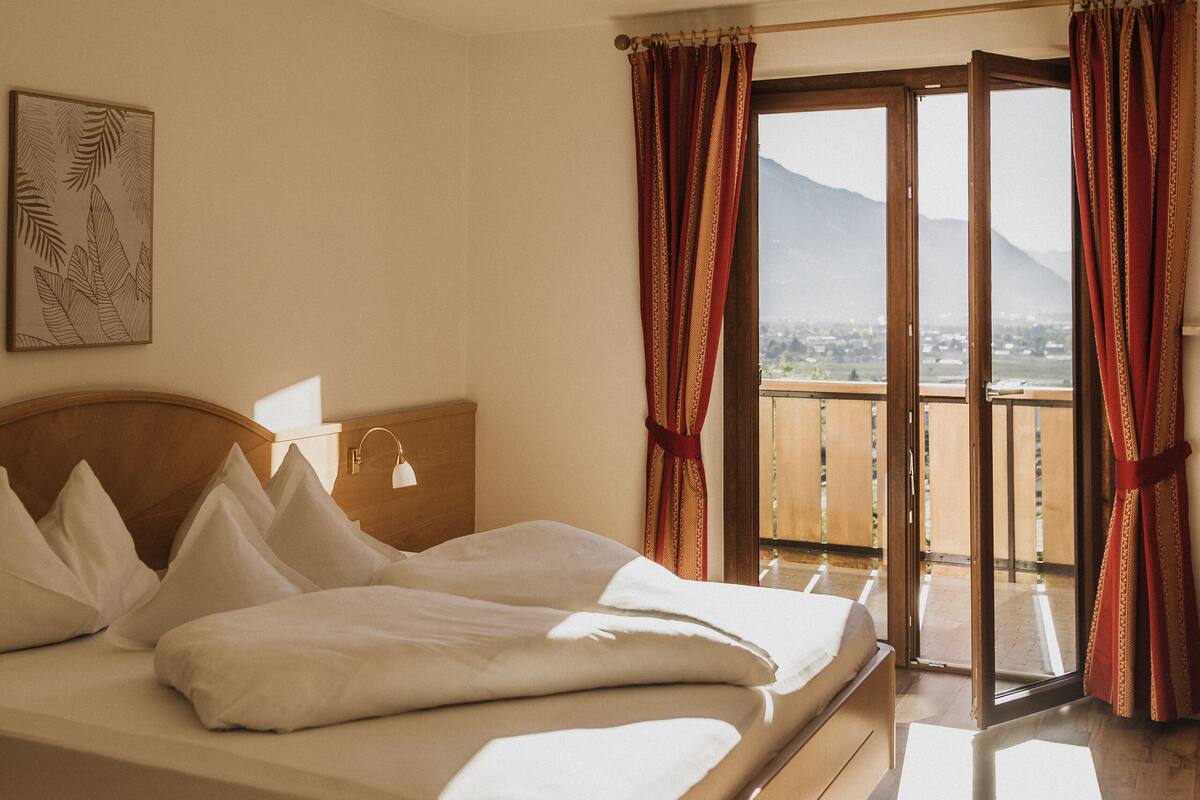 A hotel room with a bed, a bedside table, a lamp, a window with curtains, and a view of a mountain.