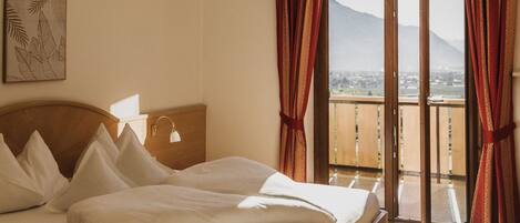 A hotel room with a bed, a bedside table, a lamp, a window with curtains, and a view of a mountain.