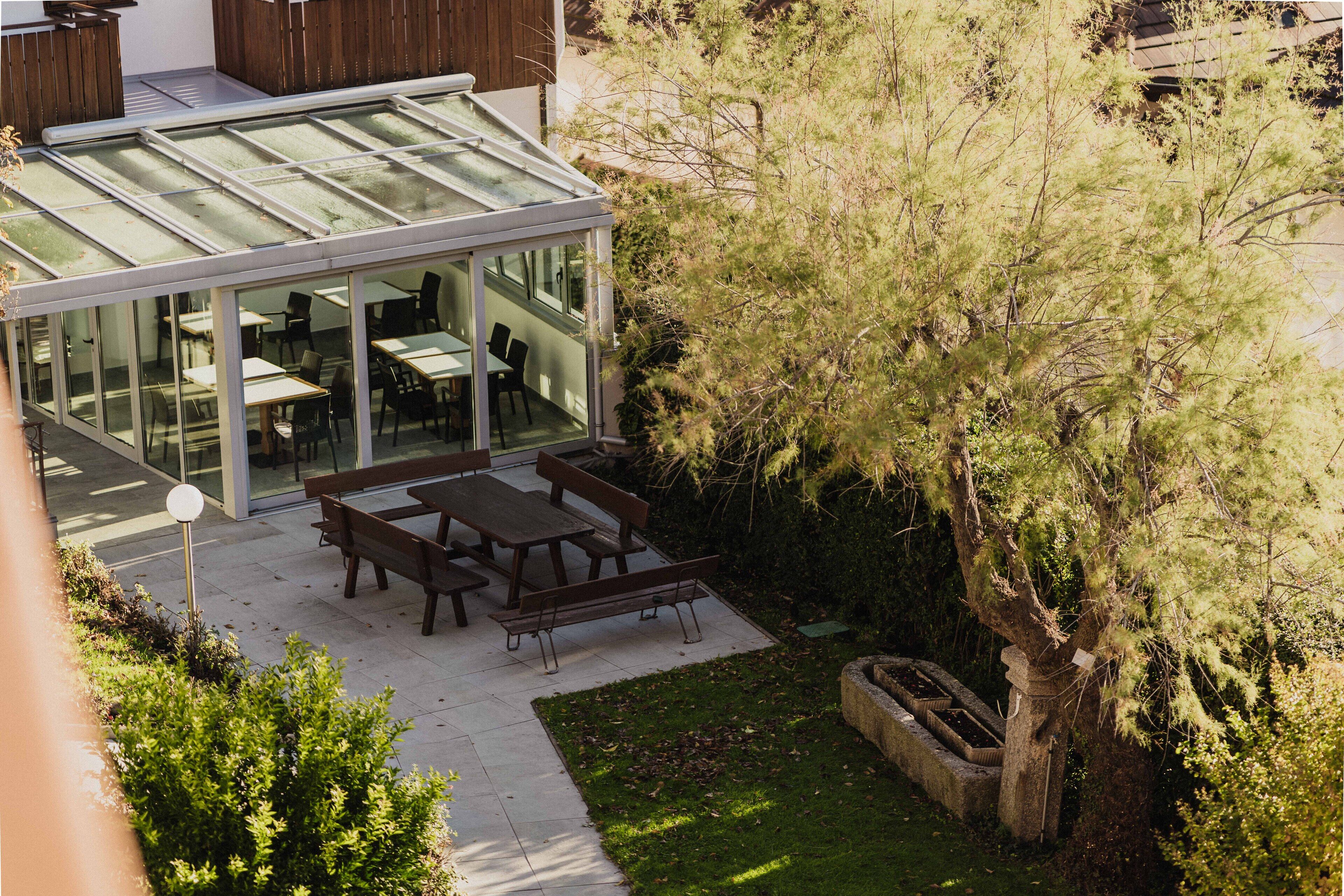 A modern outdoor dining area with glass walls, wooden tables, and benches, surrounded by greenery and a tree.