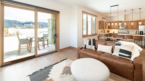 A spacious living area with a large sliding glass door leading to a balcony, a cozy brown leather sofa with patterned cushions, and a modern kitchen with wooden cabinets and pendant lighting. - Luxury Condo with Private Hot Tub: Alpenglow Chalet (Fraser)