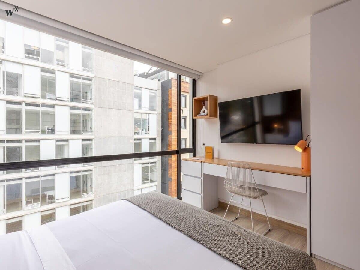 A modern hotel room with a large bed, a desk with a chair, a flat-screen TV, and a view of the cityscape through large windows.