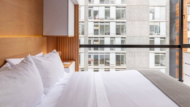 Une chambre d’hôtel moderne dotée d’un grand lit, d’une tête de lit en bois et offrant une vue sur la ville grâce à de grandes fenêtres du sol au plafond.