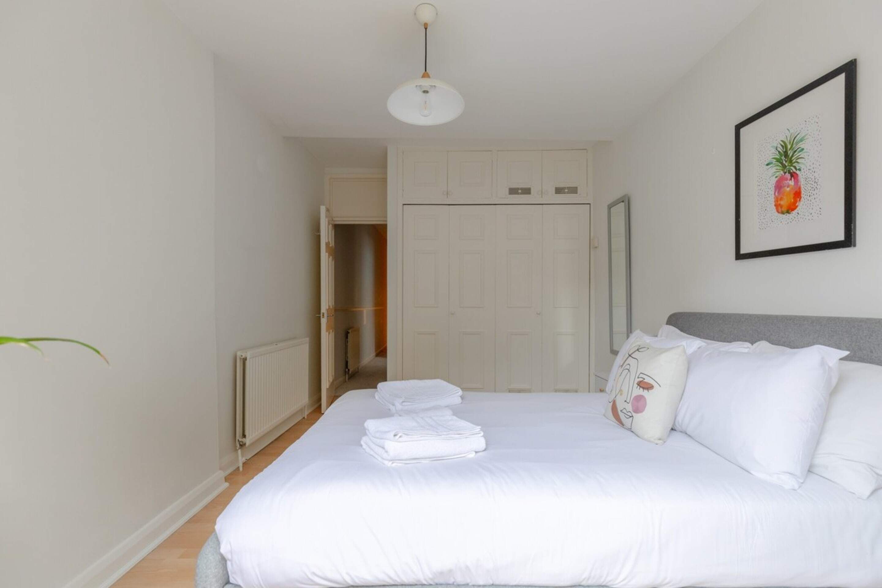 Een net opgemaakt bed met witte lakens en kussens, een witte radiator, een witte kledingkast en een afbeelding van een ananas in een lijst aan de muur.