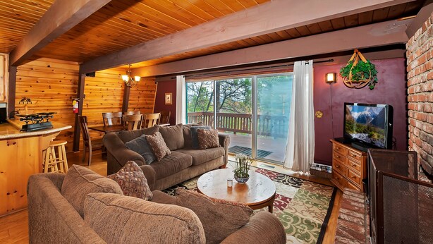 A cozy cabin interior with a wooden ceiling, a dining area with a table and chairs, a living room with sofas and a coffee table, and a TV on a wooden stand. - Tree Top Villa By Destination Big Bear:Upper Moonridge! Decks! Pool/Ping Pong Table! Foosball! (Big Bear City)