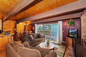 A cozy cabin interior with a wooden ceiling, a dining area with a table and chairs, a living room with sofas and a coffee table, and a TV on a wooden stand. - Tree Top Villa By Destination Big Bear:Upper Moonridge! Decks! Pool/Ping Pong Table! Foosball! (Big Bear City)