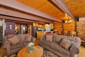 A cozy cabin interior with two sofas, a coffee table, and a kitchen area. - Tree Top Villa By Destination Big Bear:Upper Moonridge! Decks! Pool/Ping Pong Table! Foosball! (Big Bear City)
