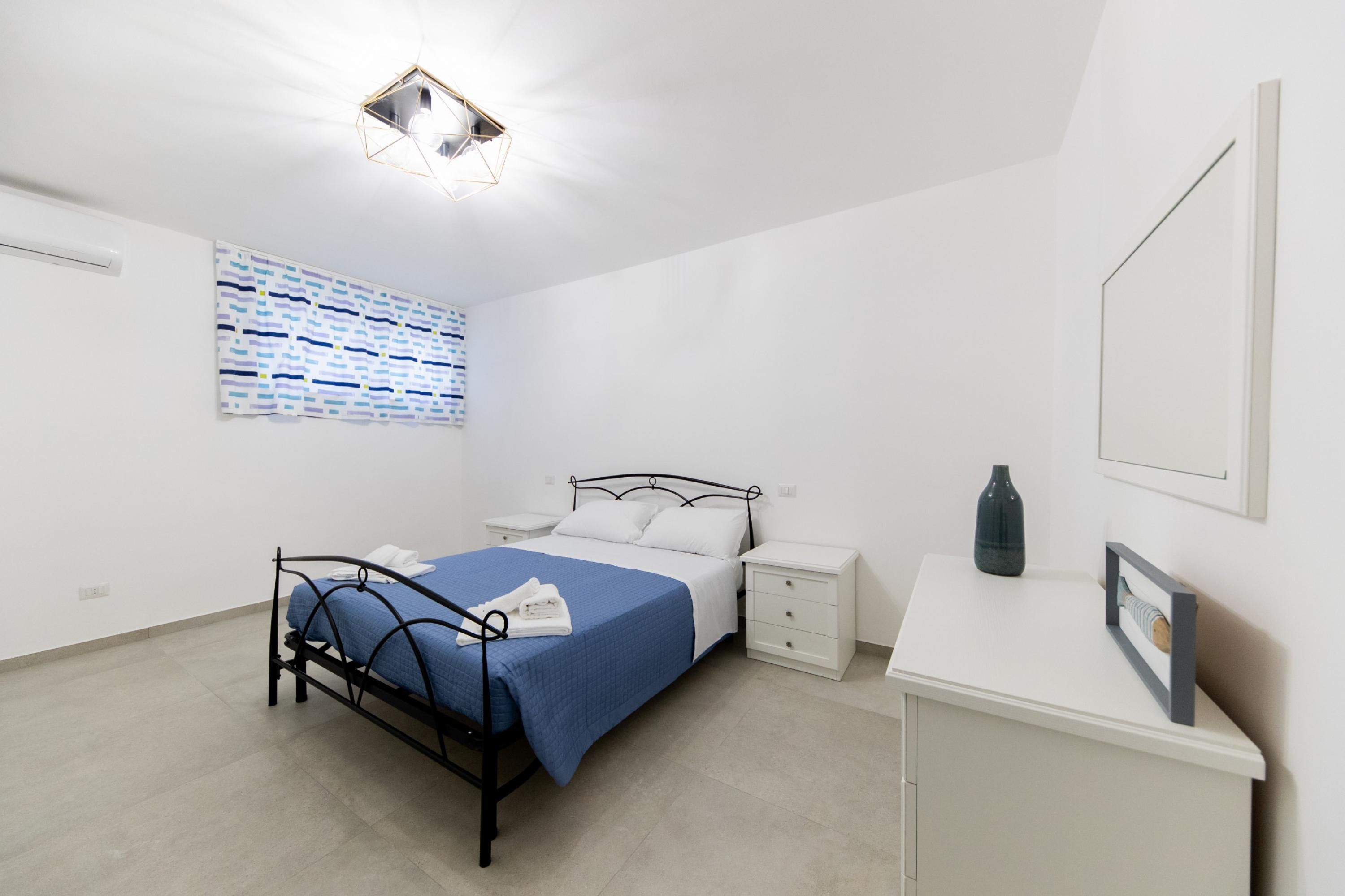 A bedroom with a bed, bedside tables, a desk, a mirror, and a geometric light fixture.