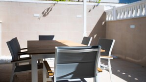 Een terras buiten met een houten tafel en stoelen, een witte balustrade en een beige muur.