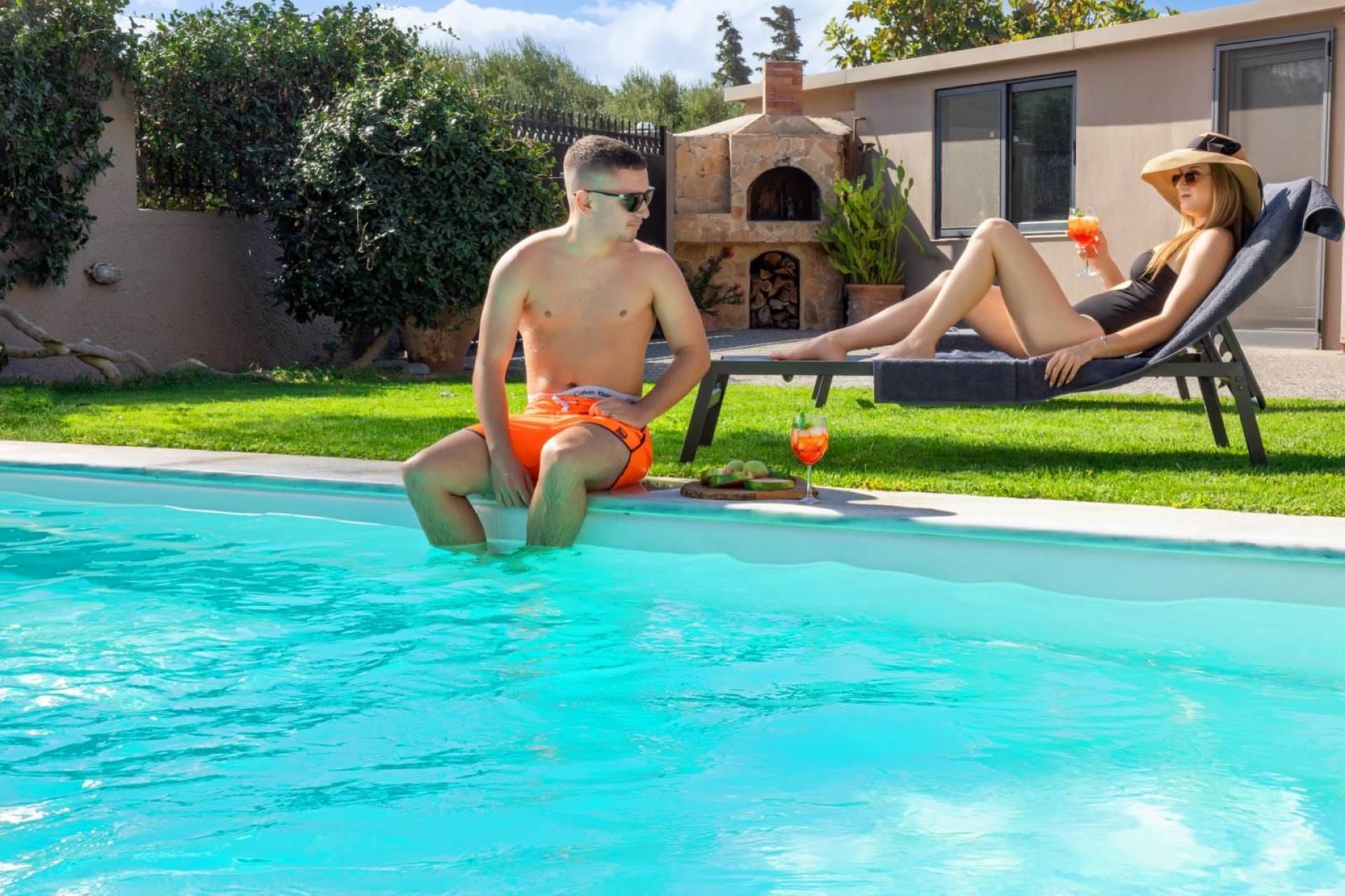A swimming pool with two people sitting on lounge chairs, a stone building with a chimney, and a clear sky.
