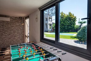 Een kamer met een tafelvoetbaltafel, een raam met uitzicht op een zwembad en gebouwen, en een stenen muur.