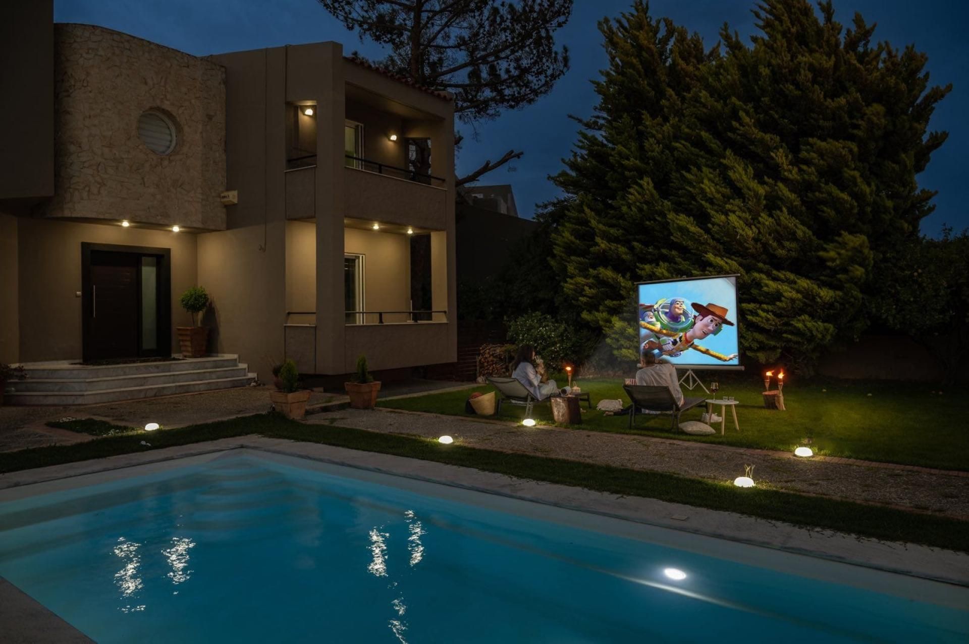 A modern house with a swimming pool, outdoor seating, and a large screen displaying a movie.