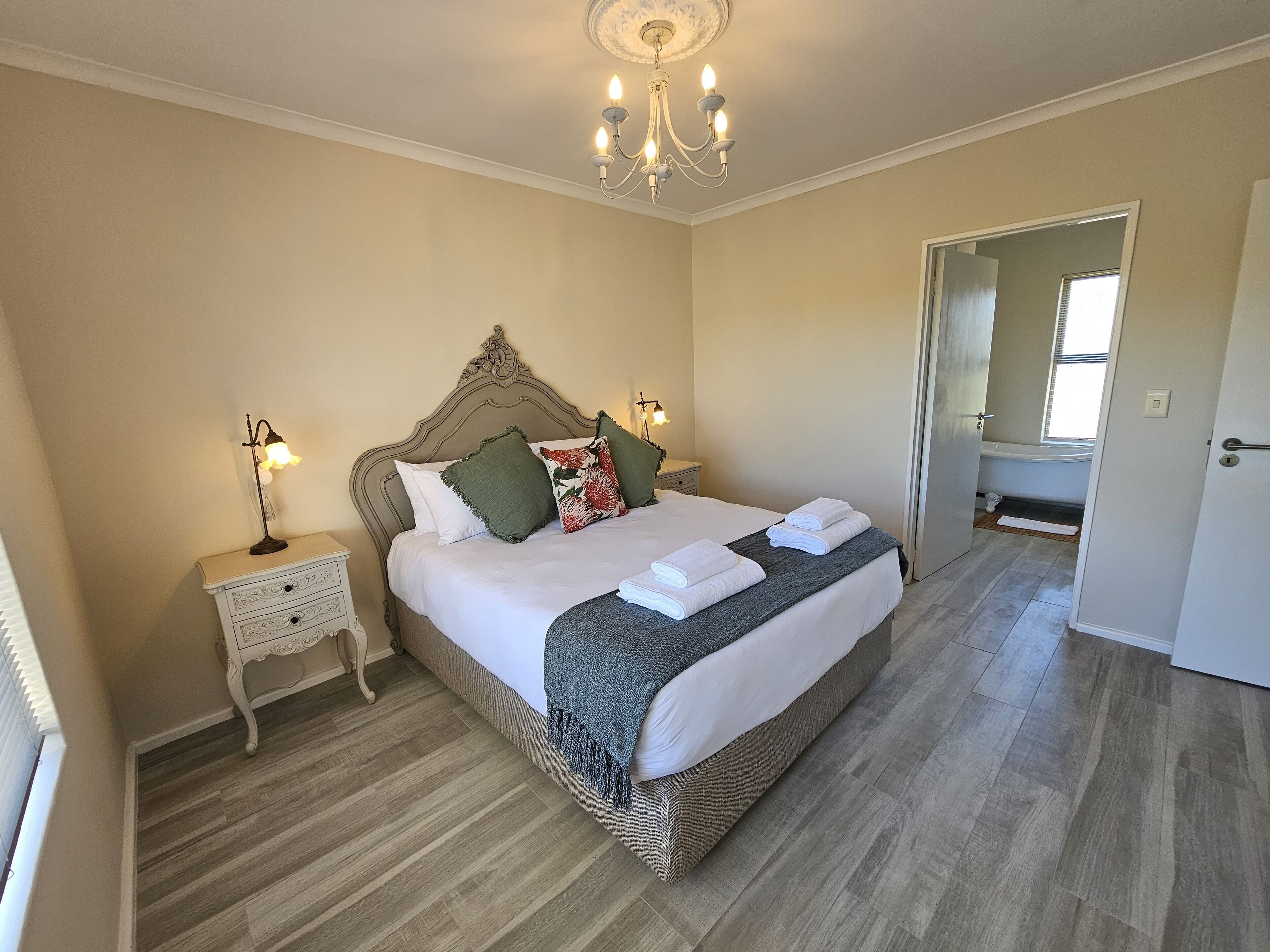 A bedroom with a large bed, a nightstand, a lamp, a chandelier, and a bathroom visible through an open door.