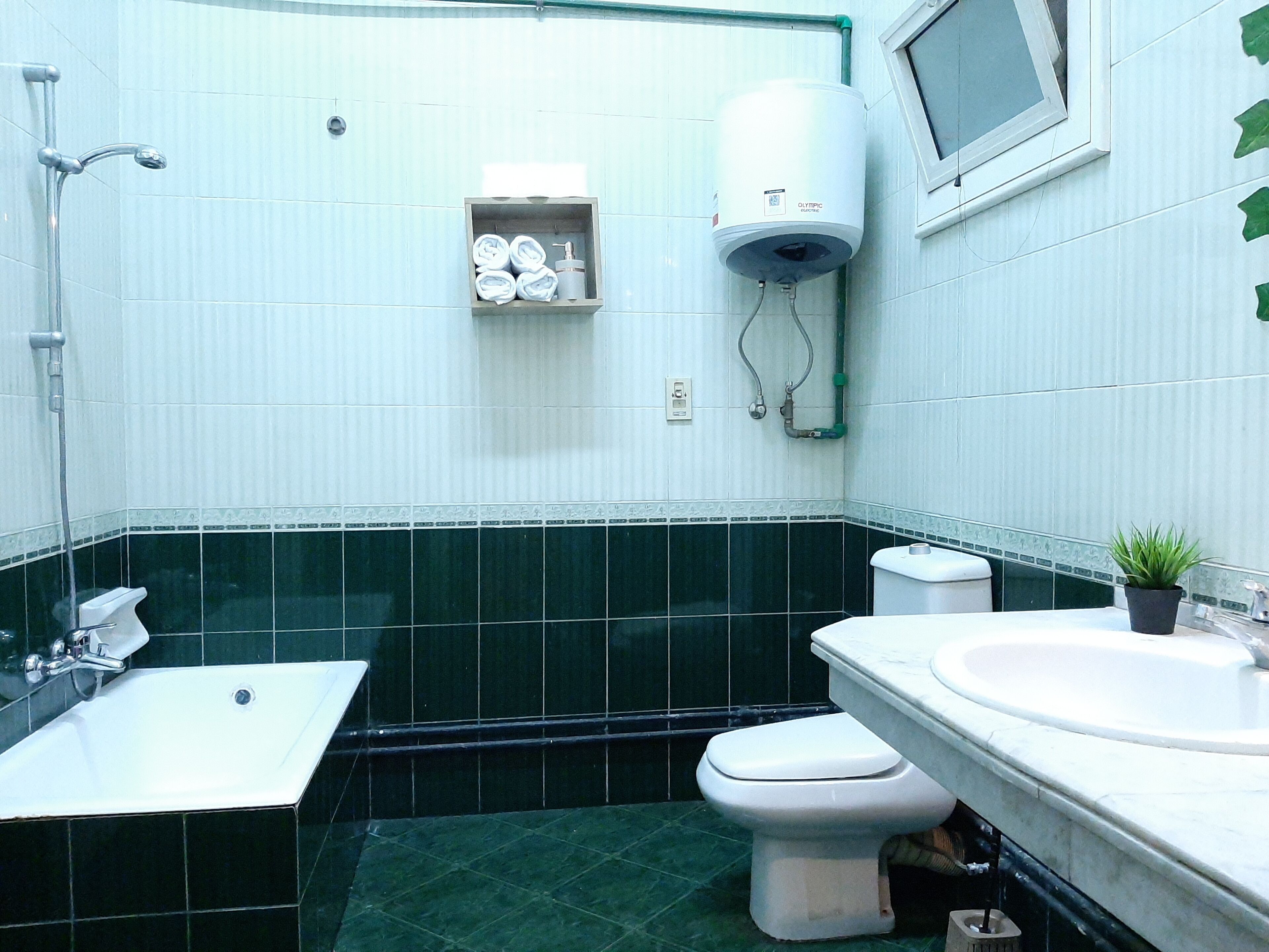 Une salle de bain avec un lavabo blanc, des toilettes et une douche. Il y a une étagère avec des serviettes et un chauffe-eau fixé au mur.