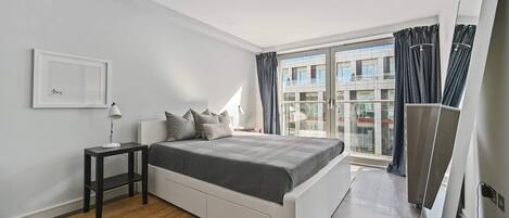 A modern bedroom with a large bed, a small bedside table, a window with curtains, and a view of buildings outside.