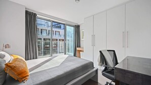 A modern bedroom with a large bed, a desk, and a view of a building through the window. - Sleek 2BD Flat W/terrace - 2 Mins From Farringdon! (London)