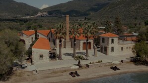 A coastal building with stone walls and red-tiled roofs, surrounded by palm trees and a beach. - Zaira Hotel (Lesvos)