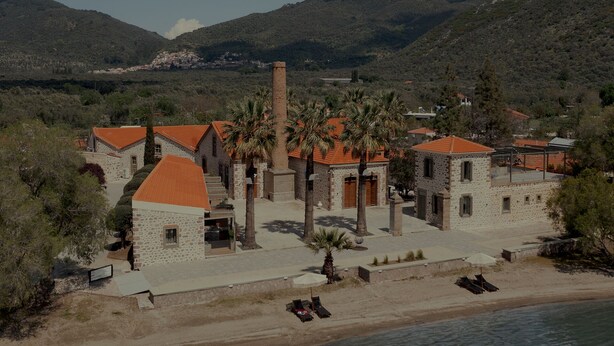 Un bâtiment situé sur la côte, avec des murs en pierre et des toits recouverts de tuiles rouges, entouré de palmiers et bordé par une plage.