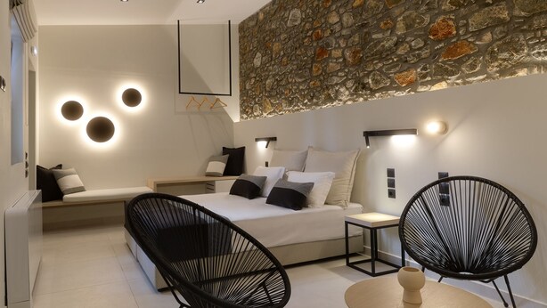 A modern bedroom with a stone accent wall, a bed with multiple pillows, a bench, two black chairs, and a small table. - Zaira Hotel (Lesvos)