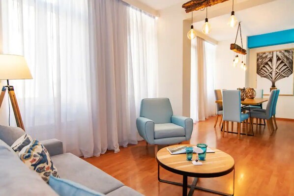 A modern living room with a sofa, armchair, and round coffee table. There is a dining area with a table and chairs in the background.