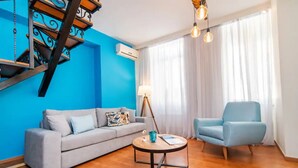 A living room with a blue wall, a grey sofa, a blue armchair, a round coffee table, and a staircase with a black railing. - Tinos La Belle vie  (Tinos)