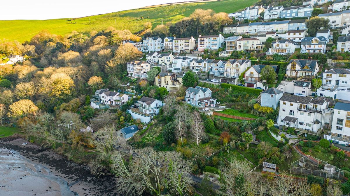 A residential area with houses on a hillside, a beach in the foreground, and a mix of greenery and buildings.