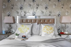 A bedroom with a tufted headboard, floral patterned wallpaper, two bedside tables with lamps, a bedside plant, and a book on the bed.