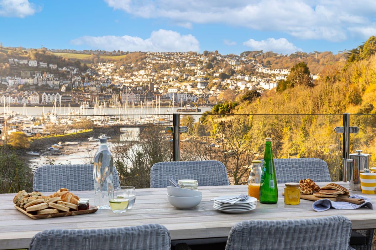 A dining table with a view of a town and marina, wicker chairs, and breakfast items.