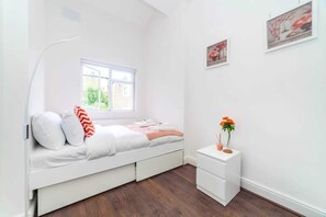 A small, white bedroom with a bed, a bedside table with a plant, and a window. - Radiant and Roomy 2BD Flat - Fulham! (London)