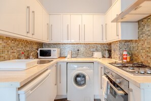 A compact kitchen with white cabinets, a washing machine, a microwave, a gas stove, and a sink. - Radiant and Roomy 2BD Flat - Fulham! (London)