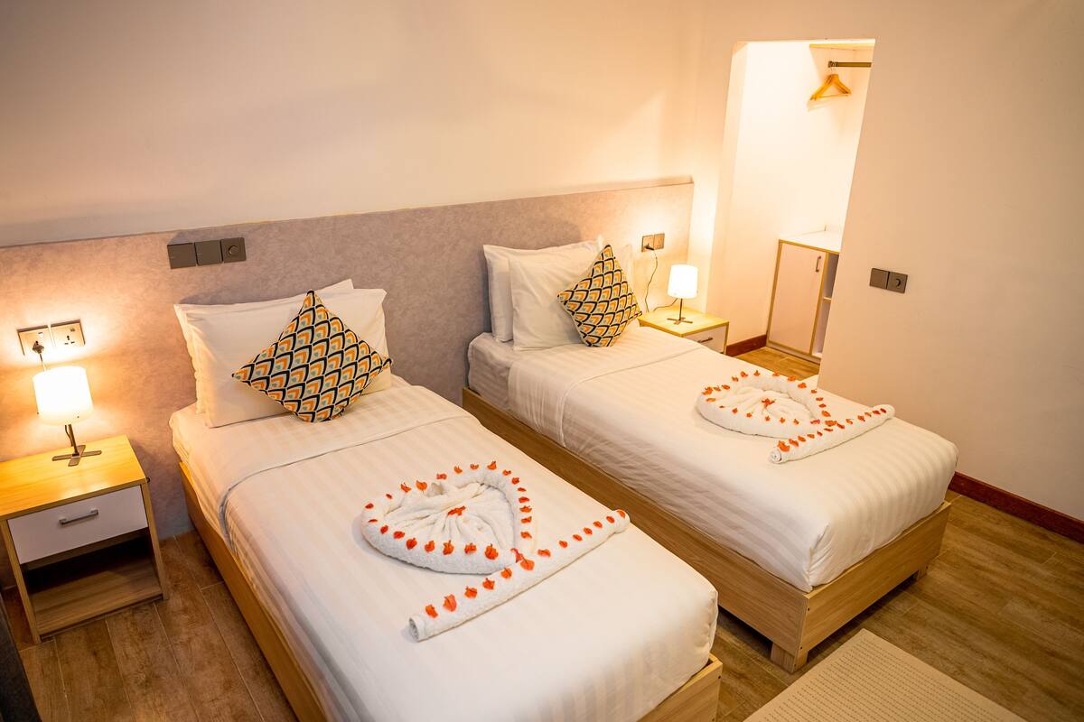 Two single beds with white linens, patterned pillows, and a towel with orange flowers. Each bed has a bedside table with a lamp.