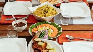 A dining table set with various dishes, including rice, fish, and vegetables, with a wooden chair and a patterned cushion in the background. - Holiday Crib (Dhiffushi)