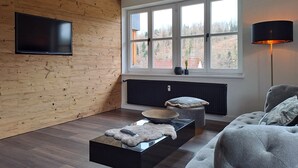 Moderni olohuone, jossa on puuseinä, suuri ikkuna, litteänäyttöinen televisio, musta sohvapöytä, harmaa sohva ja lattiavalaisin.