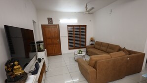 A living room with a brown sofa, a flat-screen TV, a wooden door, and a window with blinds. - Guest West Inn (Kanayannur)