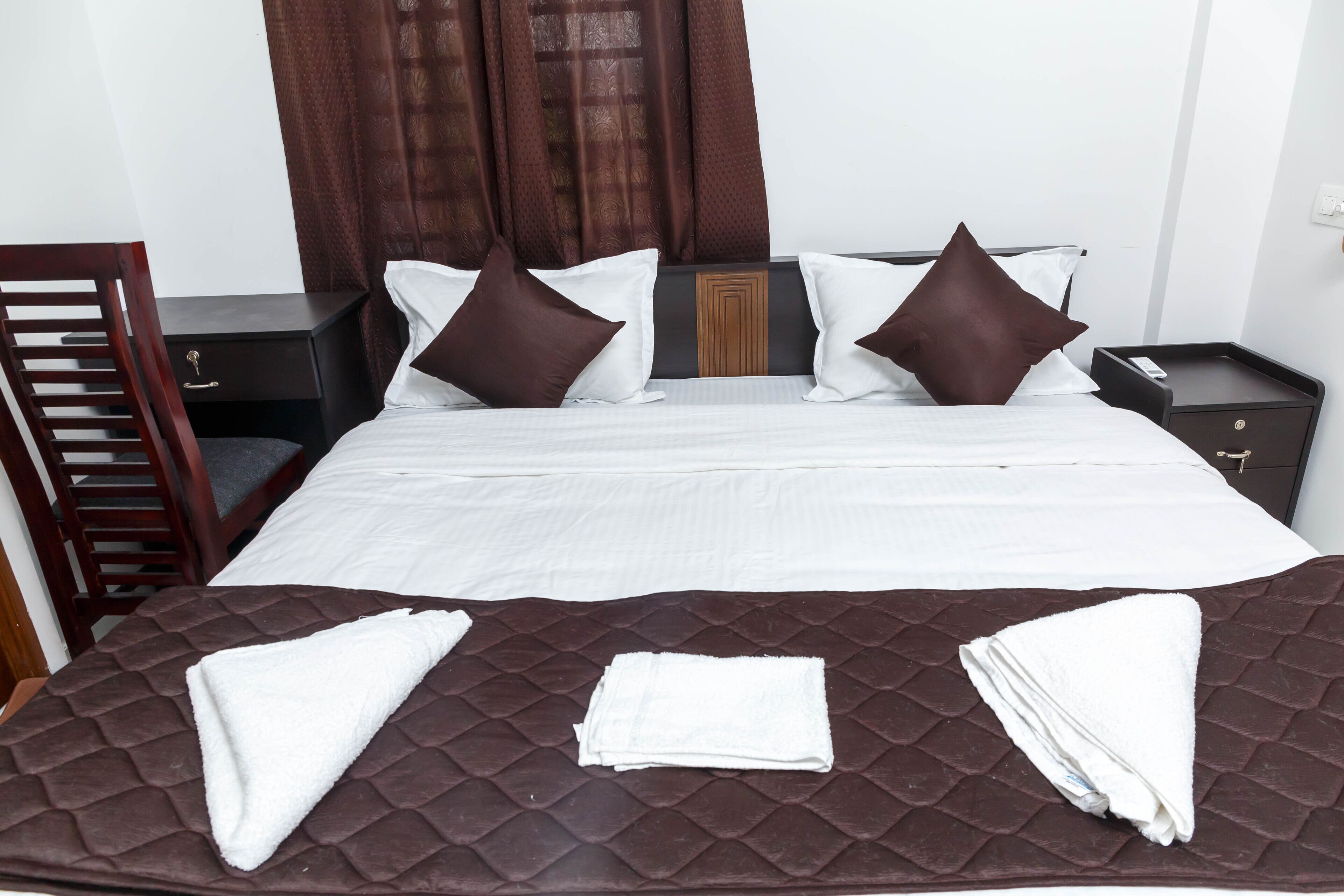 Una cama bien hecha con una cubierta acolchada marrón, dos almohadas blancas y dos almohadas marrones. Hay toallas blancas dobladas sobre la cama.