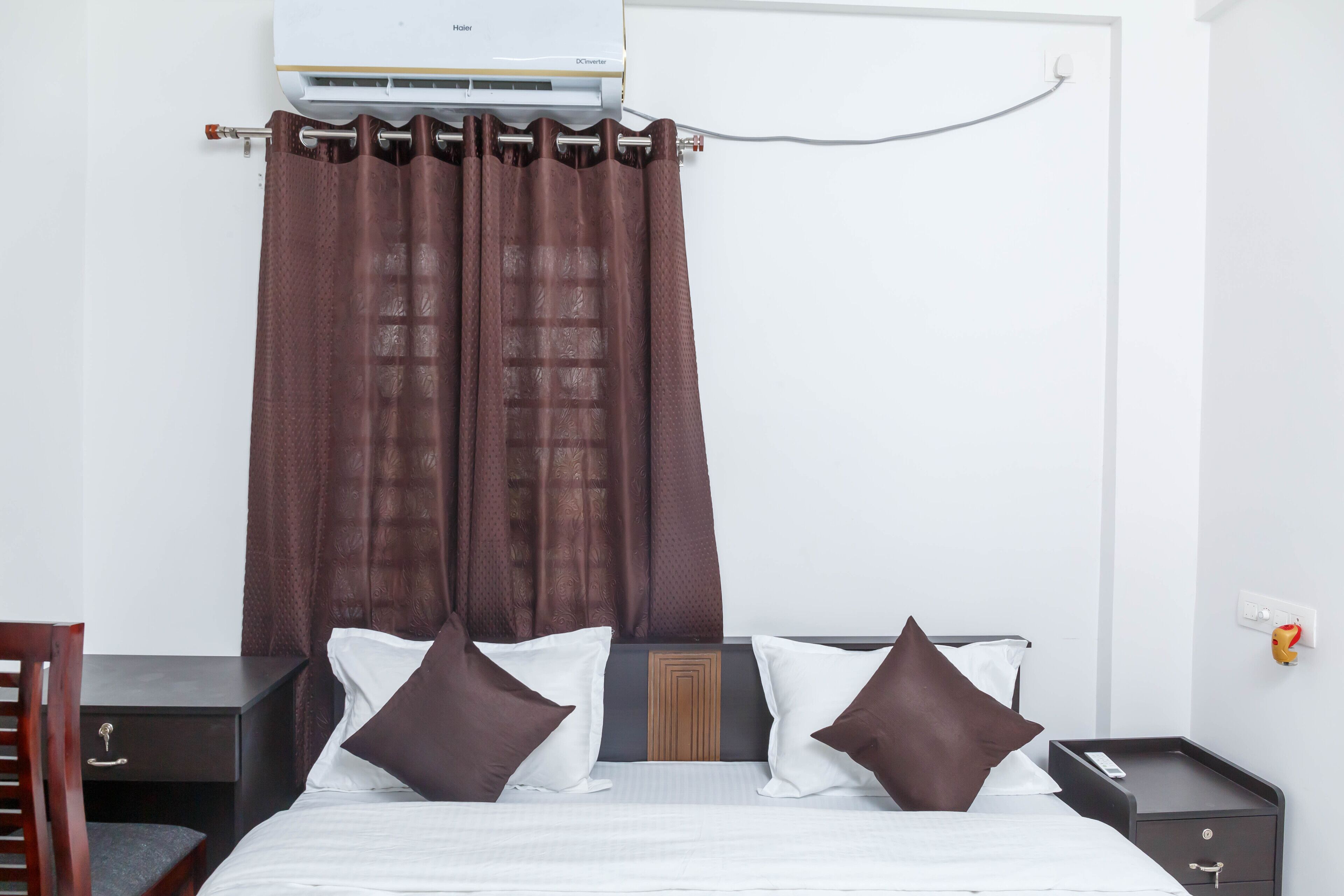 Habitación de hotel con cama, dos almohadas, mesita de noche, una silla y aire acondicionado instalado en la pared.