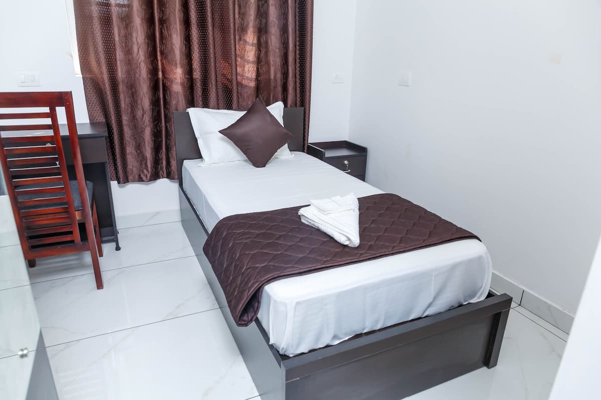 A neatly made bed with a white and brown bedspread, a wooden nightstand, a chair, and a curtain with a leaf pattern.