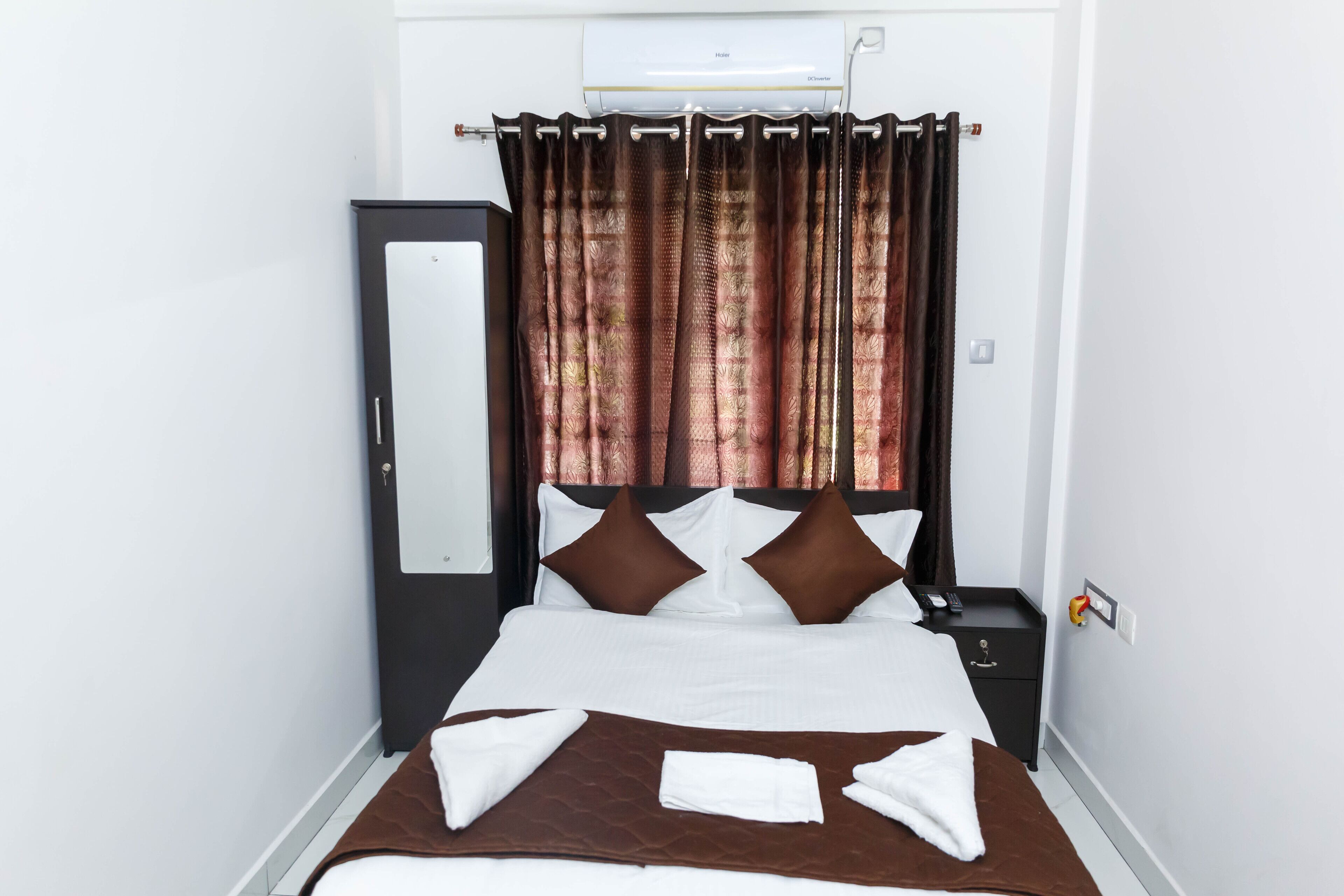 A neatly made bed with a brown bedspread, white pillows, and a brown headboard. There is a black nightstand with a remote control and a small drawer.