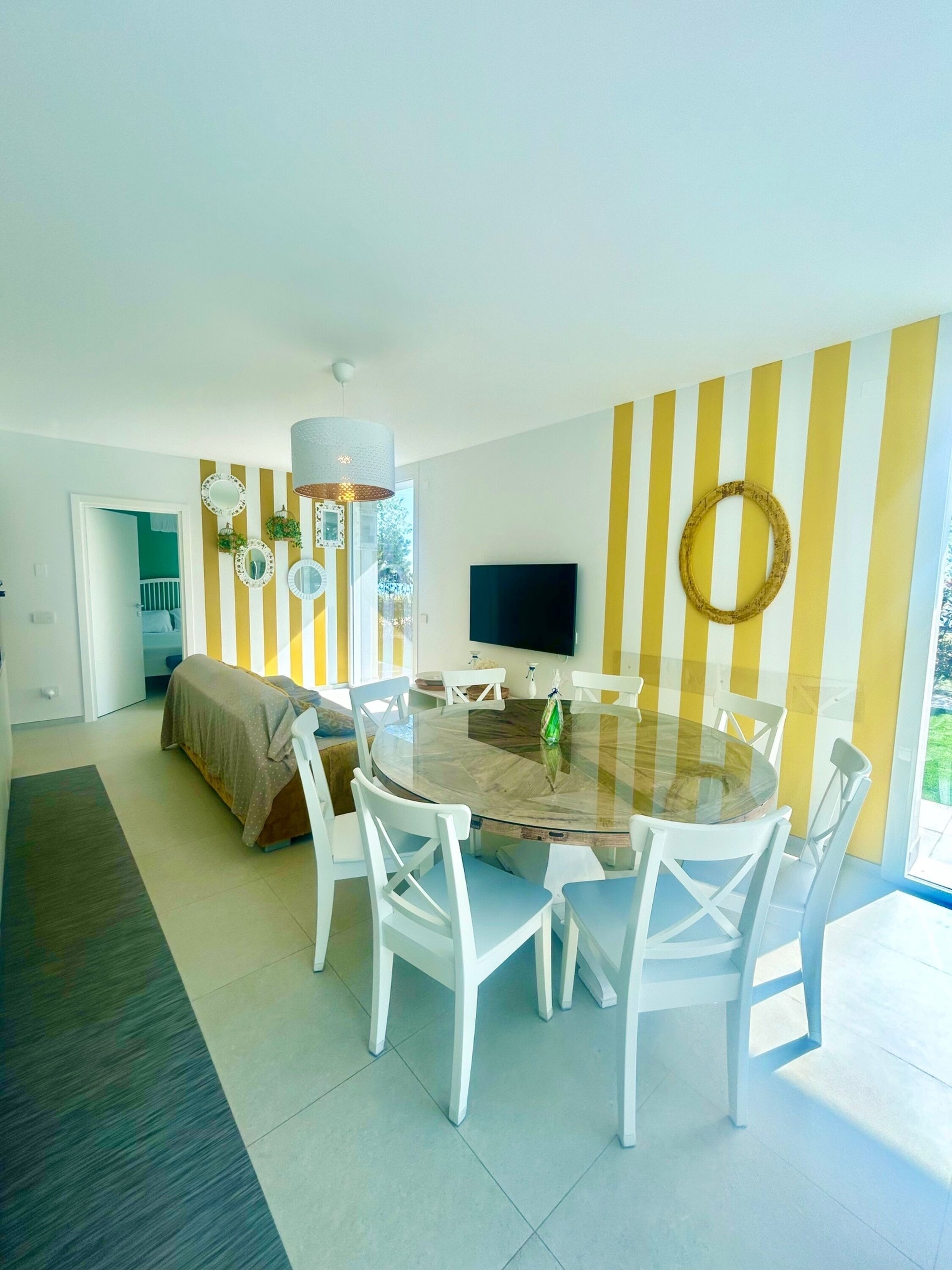 A modern living room with a round glass table, white chairs, a TV, and a yellow-striped wall.