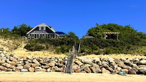 Une maison de plage avec un escalier menant à une zone sablonne, entourée de gros rochers et de verdure.