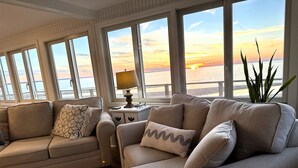 Un salon avec un canapé et des fauteuils, de grandes fenêtres offrant une vue sur le coucher de soleil sur l’eau, et une table d’appoint avec une lampe.