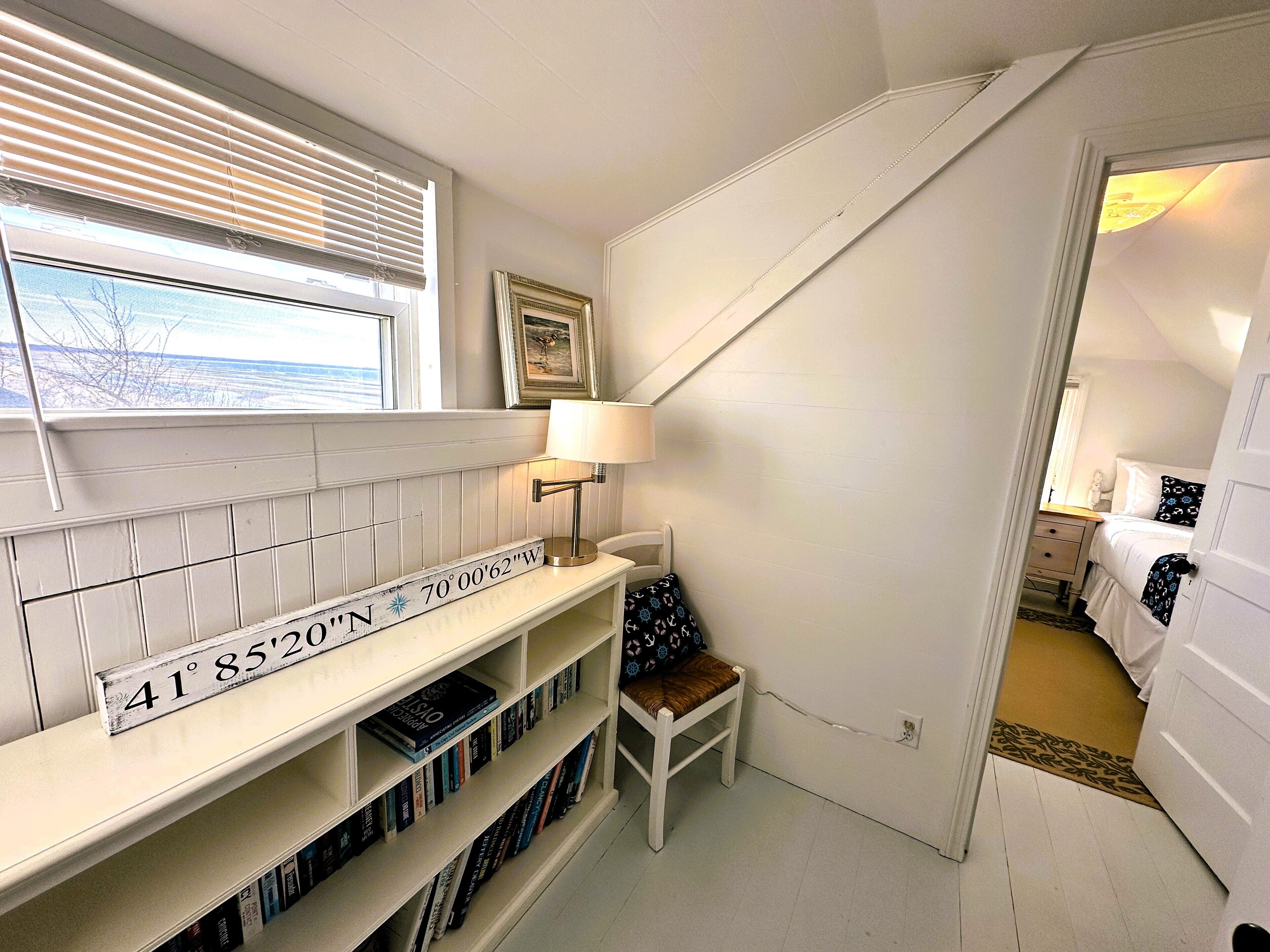 Eine Treppe mit weißer Geländer, ein Bücherregal mit Büchern, ein gerahmtes Bild an der Wand und eine Lampe auf einem kleinen Tisch.