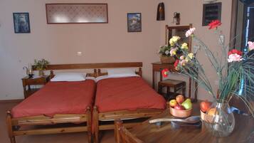 A double bed with red bedding, a wooden nightstand with a lamp, a wooden chair, a table with a fruit bowl, and a vase with flowers.