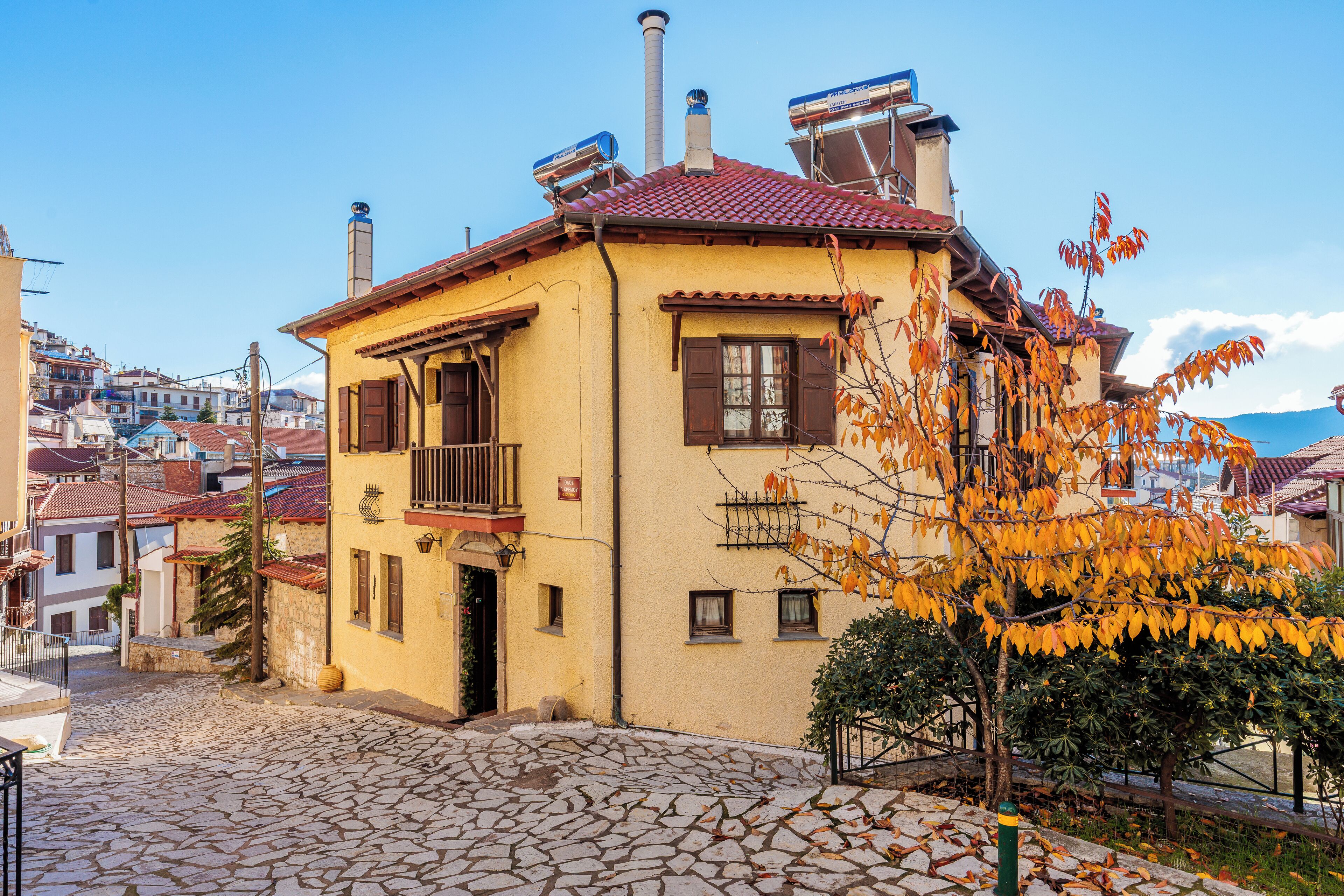 Un edificio giallo con tetto in tegole rosse, un cortile in pietra e un albero con foglie autunnali.