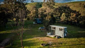 A small, modern RV-style accommodation with a deck and a hot tub in a grassy, hilly area with trees.