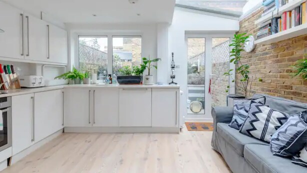 A modern kitchen with white cabinets, a central island, and a dining area with a grey sofa and patterned cushions. - Beautifully Designed 2BD House - by Queen's Park! (London)